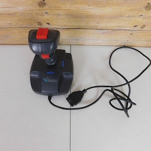 May include: A dark gray gaming joystick featuring a prominent red top button and a secondary red trigger button on the front. The base displays the Mouse Systems logo in blue and white text, and a black cable with a connector plug is attached.