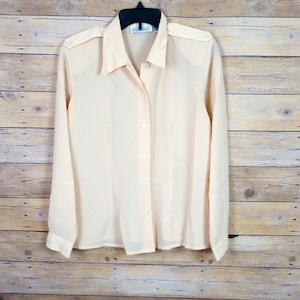 May include: A light peach-colored button-down shirt with long sleeves, a collar, and shoulder epaulets. The shirt is hanging on a black hanger against a light brown wooden background. The shirt has a classic, tailored design.