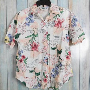 May include: White button-down shirt with a floral pattern in pink, blue, and yellow. The shirt has short sleeves and a collared neckline. The fabric also features handwritten script.