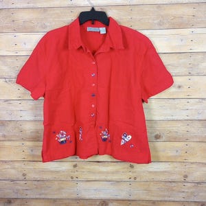 May include: A bright red short-sleeved button-down shirt with embroidered floral and heart designs. The shirt has a collared neckline and is hanging on a black hanger against a wooden background. The shirt has a label that reads "JADE MORLEY".