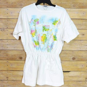 May include: A white short-sleeved t-shirt featuring a central airbrushed graphic of colorful tropical fish in shades of pink, yellow, and green against a light blue background. The garment has a gathered elastic waist and two front pockets.