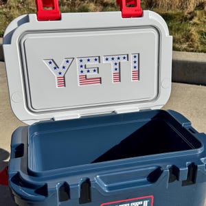May include: A navy blue hard-sided cooler with an open lid. The interior of the lid is white and features the word YETI in a blue and red American flag pattern with stars and stripes. The cooler has bright red latches at the top hinge.