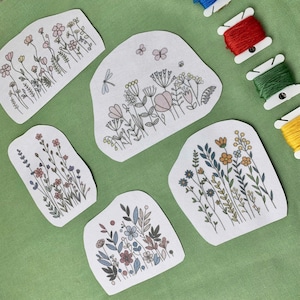 May include: Five white fabric patches featuring floral embroidery designs, including wildflowers, butterflies, and dragonflies. The patches are on a green surface, with spools of red, green, blue, and yellow embroidery thread in the background.