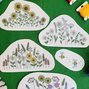 May include: A collection of embroidered patches featuring floral designs, including sunflowers, daisies, and wildflowers. The patches are in various shapes and sizes, with green, yellow, and purple accents. Two patches depict bees. The patches are on a green background.