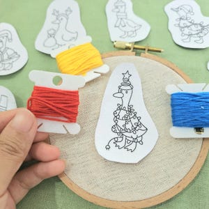 May include: An embroidery hoop with a duck design, surrounded by spools of red, yellow, and blue thread. The duck is wearing a Christmas tree outfit. The image is on a green surface.