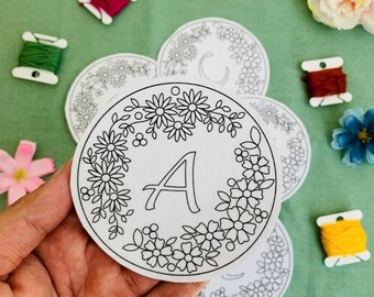 May include: A white circular coaster with a floral wreath design and the letter "A" in the center. The coaster is surrounded by other coasters, spools of thread, and flowers on a green surface.