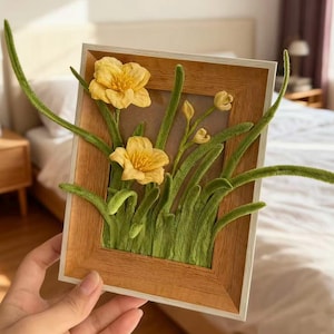 May include: A framed floral art piece featuring yellow flowers and green foliage. The flowers are the focal point, with detailed petals and a vibrant yellow hue. The frame is made of light-colored wood, adding a natural touch to the artwork.