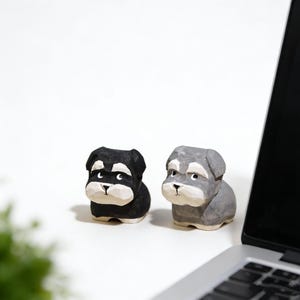 May include: Two wooden dog figurines, one black and one gray, sit side-by-side. The dogs have white accents on their faces and chests. The figurines are on a white surface, with a laptop in the background.