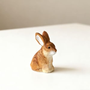 Op de afbeelding: Een kleine, handgesneden houten konijnenfiguur. Het konijn is bruin met witte accenten op de oren en snuit. Het beeldje staat op een wit oppervlak.