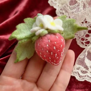 Pode incluir: Um broche de morango de feltro feito à mão. O morango vermelho tem detalhes costurados em branco, folhas verdes e uma flor branca com centro amarelo. Um acessório charmoso.