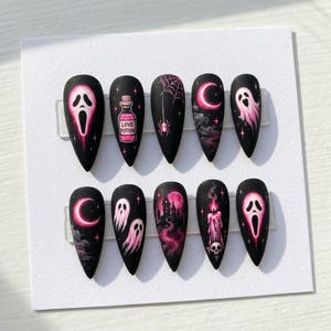 Puede incluir: Un set de diez uñas postizas almendradas con base negra mate. Los diseños pintados a mano en rosa incluyen caras de fantasmas, una botella de poción de amor, una araña con telaraña, lunas crecientes, fantasmas flotantes y una vela.