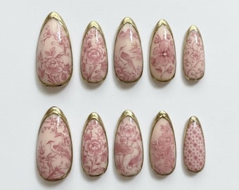 May include: Set of ten almond-shaped false nails. Each nail features a pink floral design with gold trim. The designs include flowers, birds, and other decorative elements. The nails are arranged on a white surface.