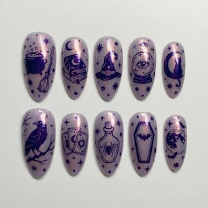 Puede incluir: Un conjunto de diez uñas postizas en forma de almendra de color lavanda. Cada uña presenta un diseño morado oscuro con temática de brujas, que incluye un caldero, un sombrero de bruja, una bola de cristal y un cuervo. Las uñas están decoradas con estrellas.