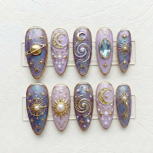 Puede incluir: Un conjunto de diez uñas artificiales con temática celestial. Cada uña presenta un diseño diferente, incluyendo planetas, lunas, estrellas y galaxias arremolinadas, en tonos morado, dorado y azul. Las uñas son de forma ovalada y tienen un acabado brillante.