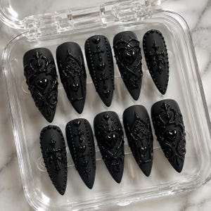 May include: A set of ten almond-shaped press-on nails with a matte black base and glossy black 3D relief designs. The raised patterns include intricate filigree, hearts, crosses, and arched window motifs, all arranged in a clear plastic storage case.