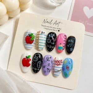 Könnte beinhalten: Ein Set aus zehn künstlichen Fingernägeln mit schulbezogenen Motiven, darunter ein roter Apfel mit grünem Wurm, Notizbuchpapier mit einer A+-Note, chemische Formeln, ein rosa Bleistift, mathematische Gleichungen, Papierflieger und bunte Büroklammern.