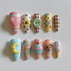 May include: A set of ten artificial nails with various designs. The nails feature a heart, flower, button, plaid, moon, floral pattern, cupcake, teddy bear, and star. Colors include pink, yellow, blue, brown, and white. The nails are oval-shaped.