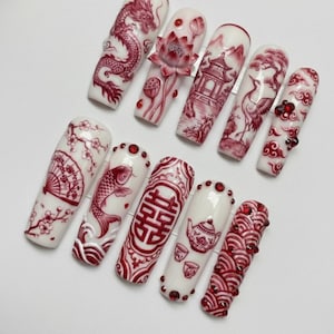 May include: A set of ten long, white artificial nails with intricate red designs. Each nail features a different Asian-inspired motif, including a dragon, lotus flowers, pagodas, and koi fish. Some nails are adorned with red gemstones.