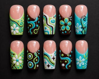 Op de afbeelding: Een set van tien kunstnagels met een natuurlijke roze basis en diverse kleurrijke ontwerpen. De ontwerpen omvatten bloempatronen, wervelingen en geometrische vormen in groen, blauw, bruin en wit, tegen een zwarte achtergrond.