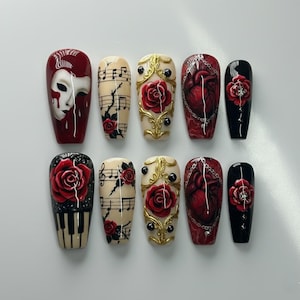 Puede incluir: Un conjunto de diez uñas artificiales con varios diseños. Los diseños incluyen una rosa roja, un teclado de piano, notas musicales, un corazón y una máscara. Las uñas están pintadas en rojo, negro y beige con detalles dorados.