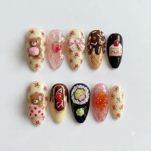 May include: A set of ten press-on nails featuring various 3D designs including a brown bear with a pink macaron, a strawberry pattern, a pink bow, an ice cream cone, a slice of cake, a bear in a strawberry blanket, chocolate, sushi, and roe.