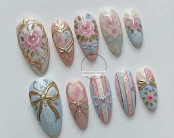 May include: A collection of ten artificial nails with various designs. The nails feature pastel pink, blue, and white colors, with gold accents, bows, and floral patterns. Some nails have striped patterns.