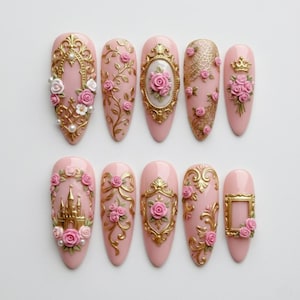 May include: A set of ten pink artificial nails with ornate gold embellishments. Each nail features a unique design, including roses, frames, and a castle. Some nails have pearl accents, creating a luxurious and detailed aesthetic. The nails are arranged on a white surface.