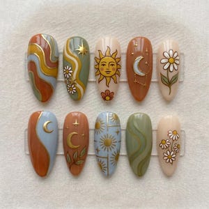 Puede incluir: Un juego de diez uñas postizas en forma de almendra con varios diseños que incluyen líneas onduladas, una cara de sol, lunas crecientes, estrellas y margaritas en una paleta de colores terracota, verde salvia, azul claro y crema con detalles dorados.