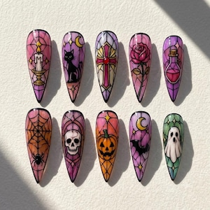 May include: A set of ten almond-shaped press-on nails featuring stained glass-style designs. The artwork includes a candle, black cat, cross, rose, potion bottle, spider web, skull, jack-o'-lantern, bat with a crescent moon, and a ghost.