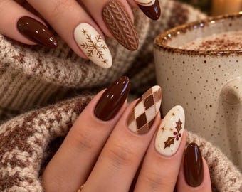 May include: Almond-shaped nails featuring a mix of solid dark brown polish, cream-colored polish with a brown snowflake design, a textured brown cable knit pattern, a brown and cream argyle print, and a cream nail with a holly berry illustration.