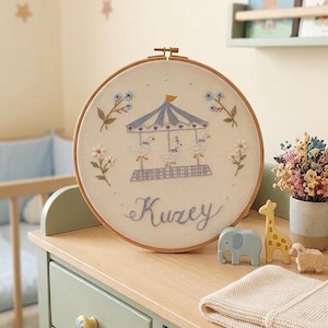 May include: Embroidered hoop art featuring a carousel design with three ducks, blue and white canopy, and the name "Kuzey" in cursive. The artwork is displayed on a light green dresser with wooden animal toys.