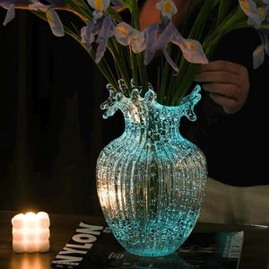 May include: A clear glass vase with a ribbed texture and a splash-like rim, illuminated with a bright turquoise glow. The vase is filled with purple and yellow iris flowers, creating a vibrant contrast against the dark, moody background.