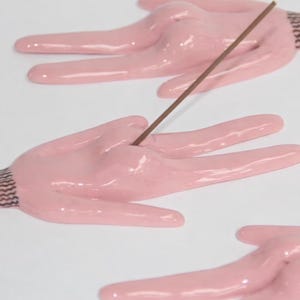Puede incluir: Soporte de incienso de cerámica rosa con forma de mano, con una varilla de incienso marrón insertada. La mano tiene una zona de muñeca texturizada. El fondo es blanco.