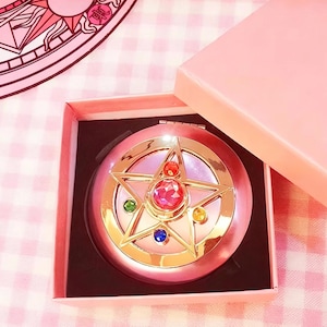 May include: A round pink compact mirror featuring a gold star emblem on the lid. The star is accented with a large central pink gemstone, a smaller red stone, and three smaller circular stones in green, blue, and yellow. It is housed in a pink box.