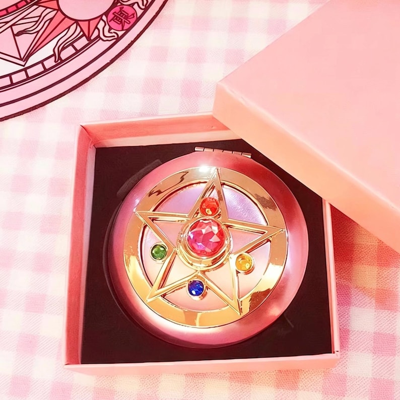 May include: A round pink compact mirror featuring a gold star emblem on the lid. The star is accented with a large central pink gemstone, a smaller red stone, and three smaller circular stones in green, blue, and yellow. It is housed in a pink box.