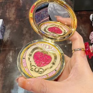 May include: A round gold-toned compact mirror with a hinged lid. The interior features a circular mirror and a decorative base plate with a central pink heart motif, gold star accents, and intricate filigree patterns surrounding the heart.
