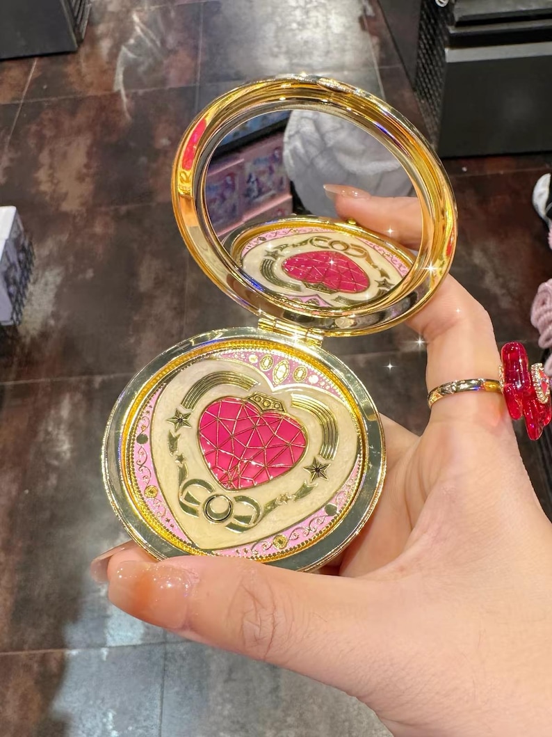 May include: A round gold-toned compact mirror with a hinged lid. The interior features a circular mirror and a decorative base plate with a central pink heart motif, gold star accents, and intricate filigree patterns surrounding the heart.