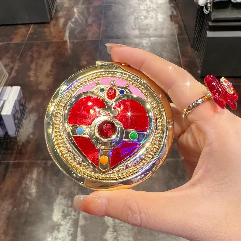 May include: A round gold compact mirror featuring a central red heart emblem adorned with small blue, green, yellow, and red gemstones. The design includes a crown-like top detail and a pink border, all set within a metallic gold frame.