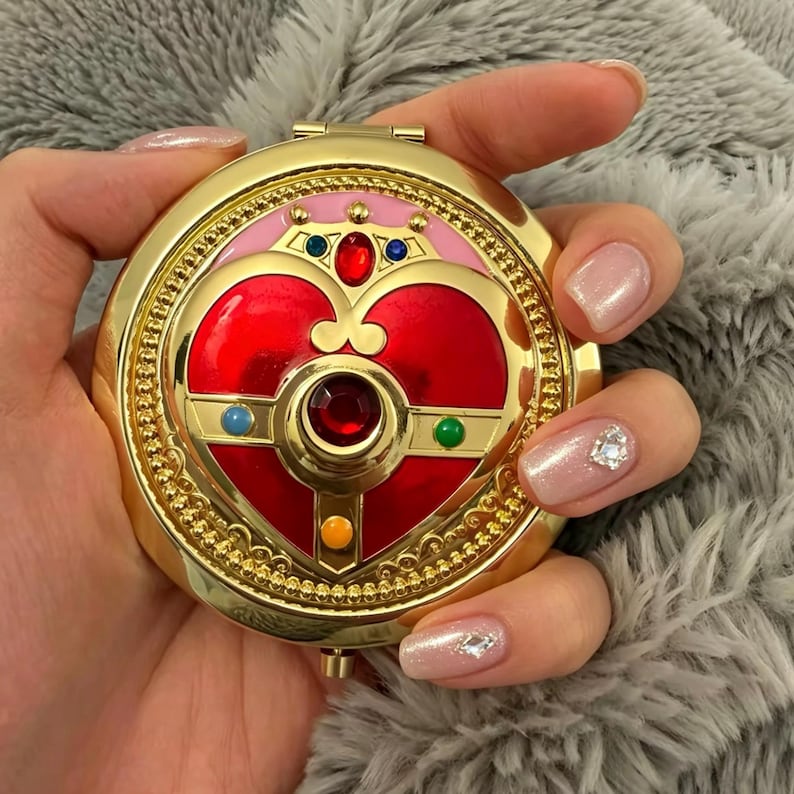 May include: A round gold compact mirror featuring a prominent red heart-shaped emblem at the center. The design includes a small crown detail, a central red gemstone, and smaller blue, green, and yellow circular accents set against a pink background.