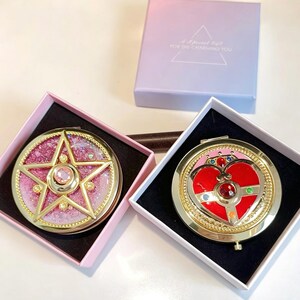 May include: Two round compact mirrors in square gift boxes. One mirror features a gold star over a pink glitter background with a central pink gem. The other mirror has a red heart design with gold accents and small colored gems. A purple box is included.