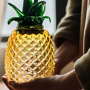May include: A decorative pineapple-shaped object crafted from translucent yellow glass with a textured, diamond-patterned surface and a vibrant green glass top featuring pointed leaves, capturing light to create a glowing, warm visual effect.