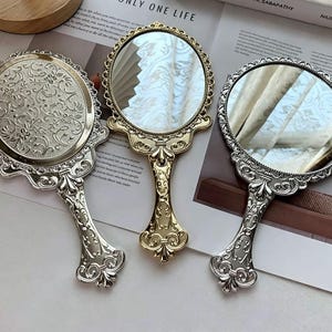 Op de afbeelding: Drie sierlijke handspiegels in goud- en zilverkleuren. De spiegels hebben decoratieve frames en handvatten met ingewikkelde bloemen- en krulwerkontwerpen. Eén spiegel heeft een effen achterkant met een bloemenpatroon, de andere hebben reflecterende oppervlakken.