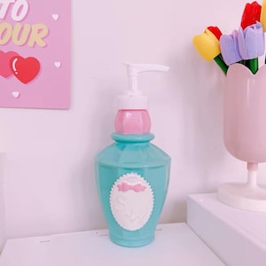 May include: A turquoise plastic soap dispenser with a white pump mechanism and a pink neck collar. The bottle features a white oval label with a small pink bow detail at the top. The design includes a faceted body and a smooth, rounded base.