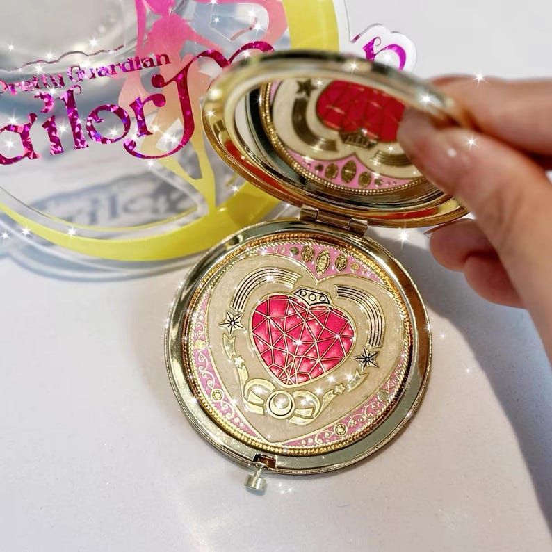 May include: A gold-toned compact mirror featuring a heart-shaped design with a faceted pink center, surrounded by cream and gold accents. The lid is open, revealing a reflective surface, and the packaging displays the text Pretty Guardian Sailor J.