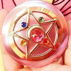 May include: A round, metallic pink compact mirror featuring a gold-toned star emblem on the lid. The star is adorned with a central faceted pink gem, surrounded by four smaller circular gems in blue, green, orange, and red, set within a gold frame.