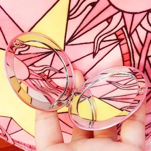 May include: A small, oval-shaped, pink-rimmed compact mirror that opens on a central hinge to reveal two reflective surfaces. The frame is metallic pink, and the mirror is positioned against a background featuring a pink and yellow sun pattern.