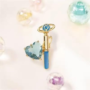Puede incluir: Broche dorado de temática fantástica, con una gema azul en forma de corazón y un diseño de varita mágica azul y dorada. El broche tiene un detalle de anillo celestial en la parte superior, sobre un fondo suave y borroso con esferas decorativas.