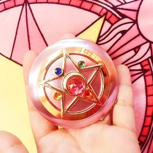 May include: A round, light pink compact mirror featuring a gold-toned star emblem on the lid. The star is adorned with a large, faceted pink center stone, surrounded by smaller, circular inset stones in blue, green, orange, and red colors.