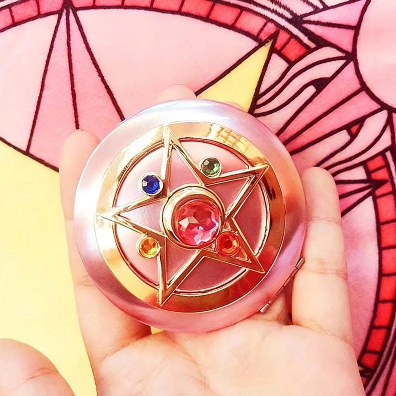 May include: A round, light pink compact mirror featuring a gold-toned star emblem on the lid. The star is adorned with a large, faceted pink center stone, surrounded by smaller, circular inset stones in blue, green, orange, and red colors.