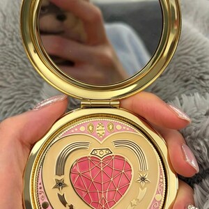 May include: A round gold-toned compact mirror featuring a hinged lid with a decorative design. The lid displays a central pink faceted heart emblem surrounded by gold star motifs, curved lines, and a pink border with small gold circular accents.